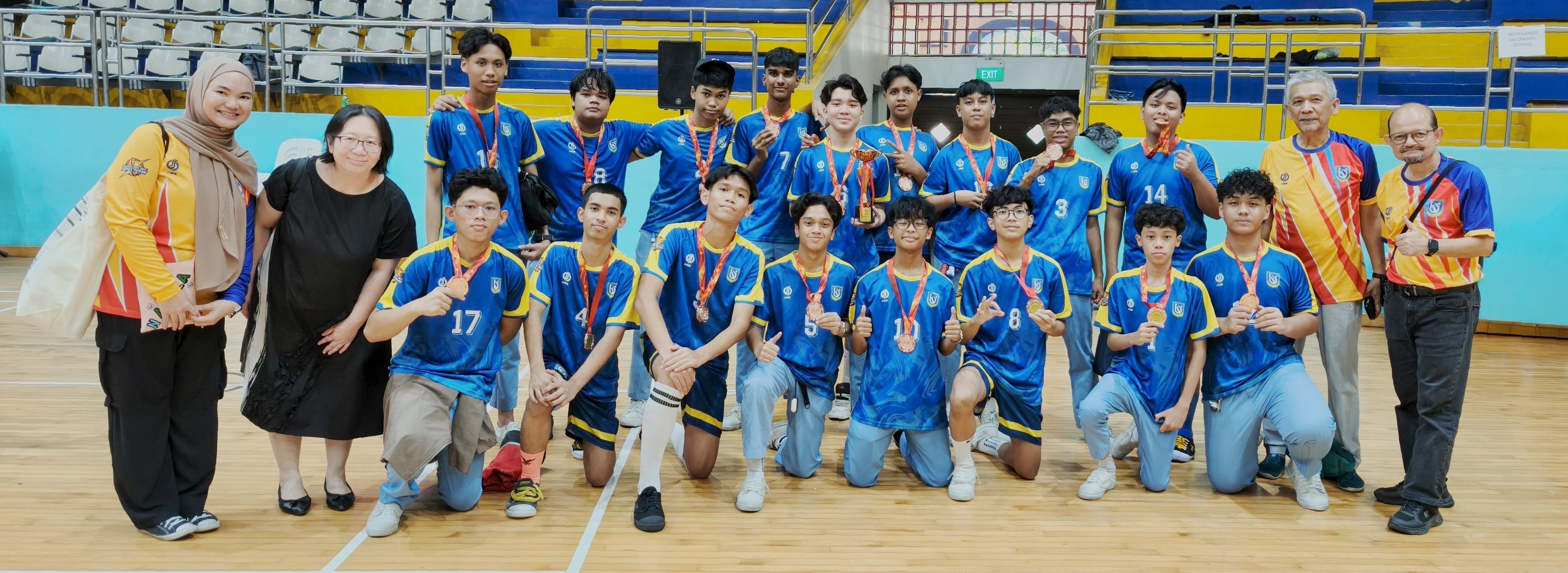 A youth sports team in blue jerseys poses with medals and a trophy alongside adults in an indoor gymnasium.